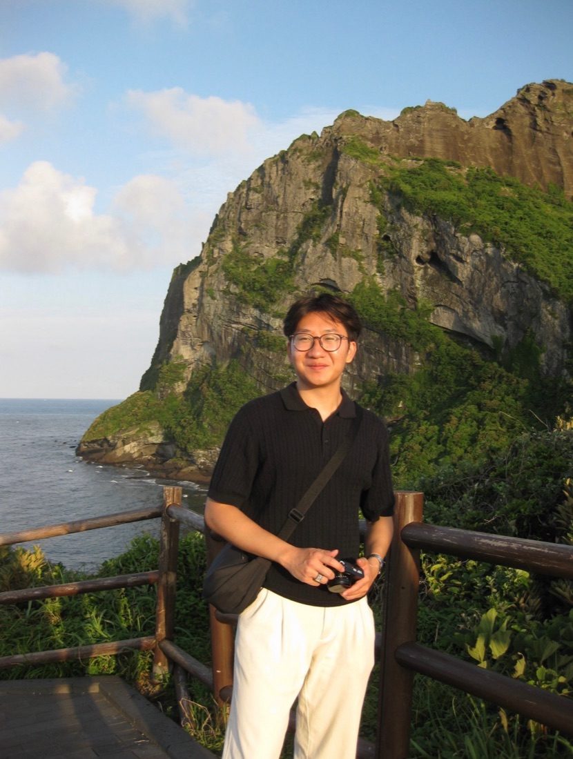 Jin holding a camera in front of a green sea cliff