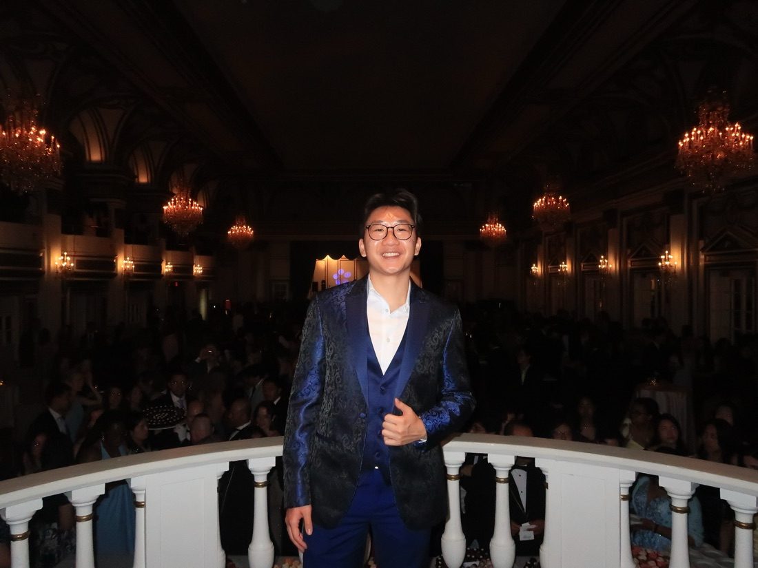 Jin smiling in a blue suit on a ballroom balcony